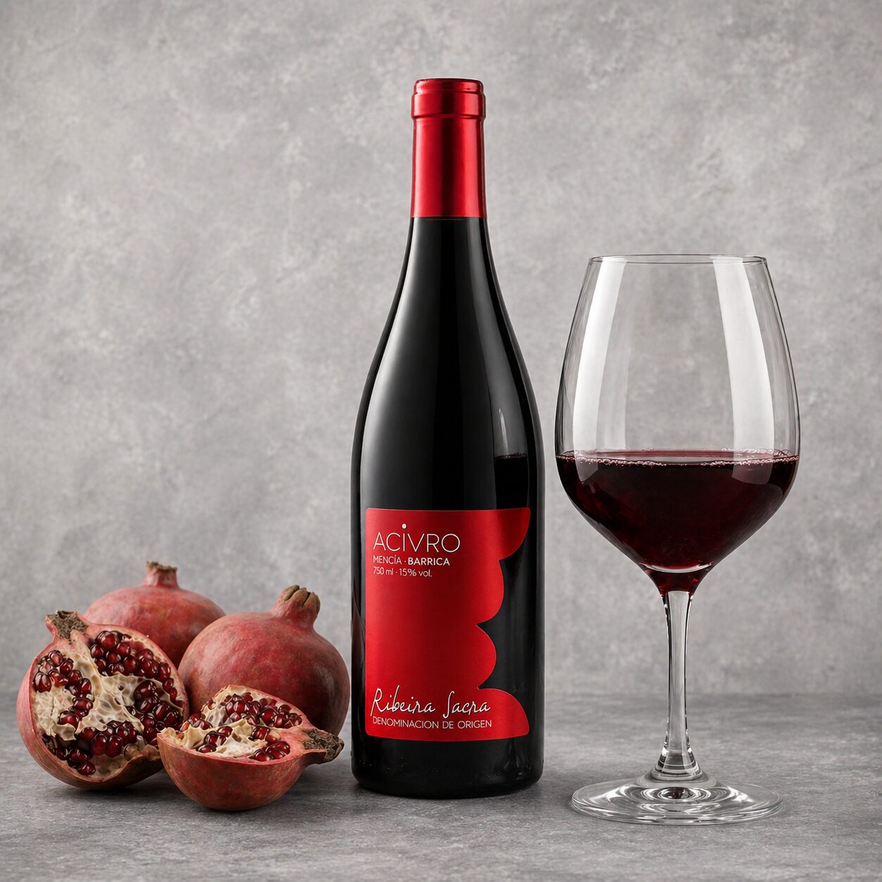 Botella de vino tinto Acivro Mencía Barrica junto a una copa de vino llena y varias granadas frescas, algunas abiertas mostrando sus semillas, sobre un fondo gris texturizado.