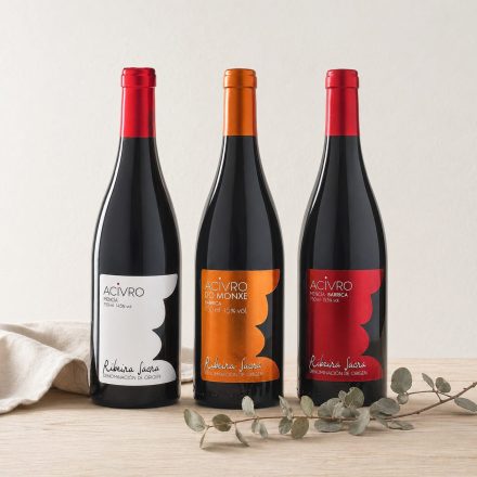 Tres botellas de vino tinto Acivro de la D.O. Ribeira Sacra (Mencía, Do Monxe y Barrica) alineadas sobre una mesa de madera con decoración minimalista.
