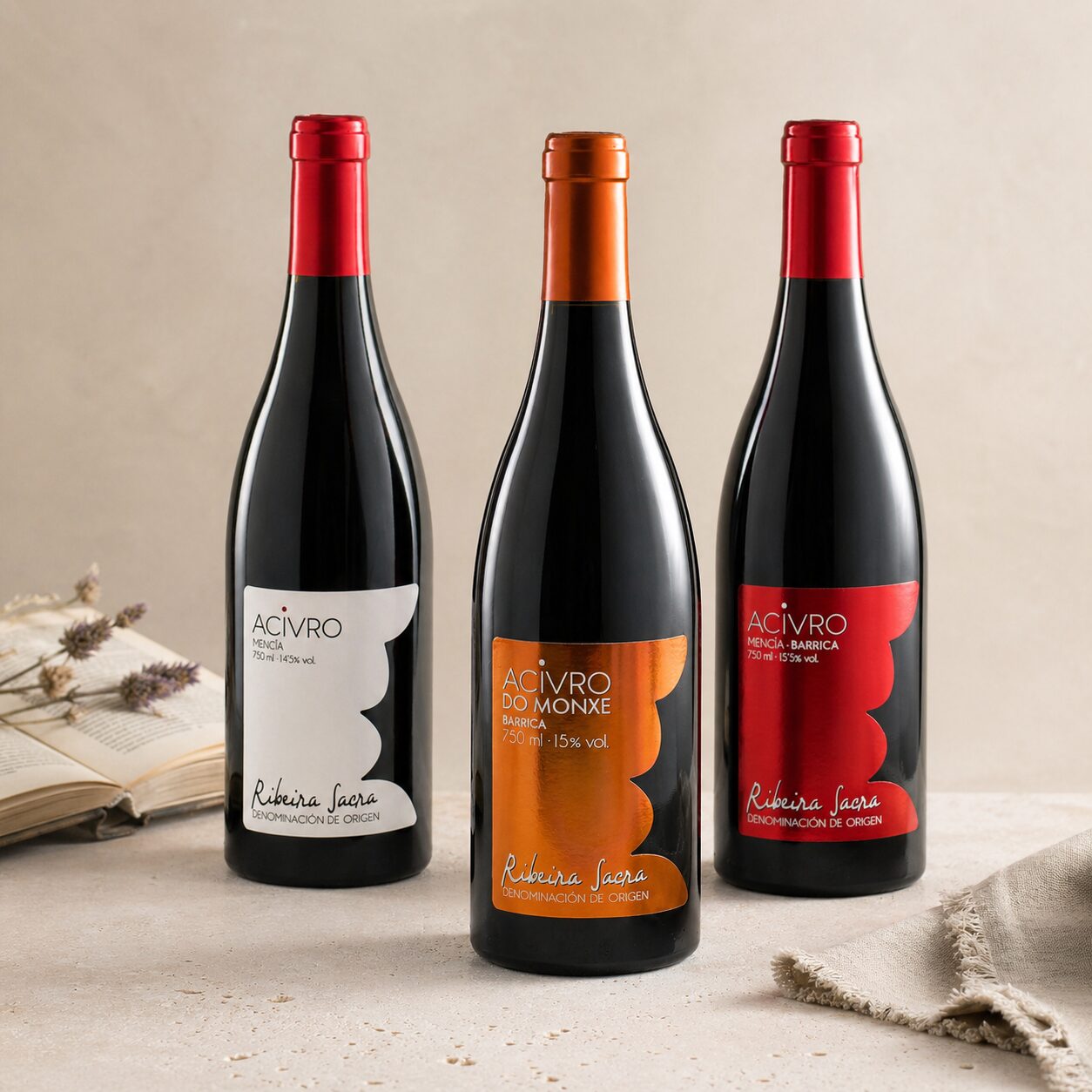 Tres botellas de vino tinto de la bodega Acivro de la D.O. Ribeira Sacra, dispuestas junto a un libro abierto, flores de lavanda seca y una servilleta de lino sobre una superficie de piedra clara.